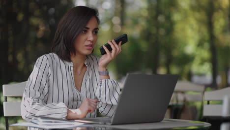 Confident-business-woman-recording-audio-message-on-smartphone-at-street.-Woman-sitting-in-a-summer-cafe-uses-smartphone-voice-recognition-send-voice-message.-speaker-or-leaving-voice-message-concept